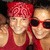 Two people in red clothing pose closely together, one wearing a red headwrap and the other with dark sunglasses on their face.