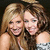 Two women with long hair pose closely together, one in a dark sleeveless top and the other in a light-colored strapless dress against a dark background.