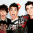Three individuals in dark clothing with red accents pose closely together against a light background, one wearing a checkered shirt.