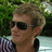 Pixelated image of a person with short light brown hair wearing dark sunglasses and a dark sleeveless top, standing outdoors with green foliage and light-colored structures in the background.