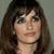 A pixelated close-up of a person with shoulder-length brown hair, dark bangs, and light skin, facing forward against a neutral gray background.