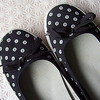 Black flat shoes with white polka dots and fabric bows on the top, positioned on a light-colored textured surface.