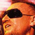 Pixelated static image of a person's face with dark hair and sunglasses, rendered in warm orange-yellow tones against a blurred background.
