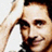 A person with dark hair and a mustache holds their right hand to their forehead in a saluting gesture, smiling slightly against a light background.