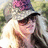 A person with long blonde hair wears a gray cap with a purple pattern and dark sunglasses, standing outdoors with green foliage in the background.