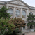 Static image of a classical building with columns, surrounded by green trees and brown foliage under a light sky.