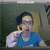 Pixelated static image of a person with dark hair and glasses wearing a blue top, holding a light-colored object near their face against a muted green background.