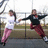 Two people in white and black tops with pink pants pose dynamically against a chain-link fence background.