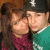 Two people in close physical proximity, one wearing a dark cap with a white emblem and teal shirt, the other in a maroon top, against a brown background.