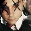 A close-up of a person with black face paint resembling spider legs over the eyes, red eyes, a nose ring, and a white collared shirt under a dark jacket.
