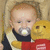 A baby with a pacifier holds a yellow teddy bear in red clothing, seated against a light brown background.