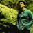 Pixelated image of a person with long dark hair wearing a teal garment, standing against a green and brown background.