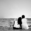 Black-and-white static image of two silhouetted figures sitting side-by-side on a beach facing the ocean, with gentle waves and an overcast sky in the background.