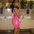 A woman in a pink dress stands in a kitchen, her arms crossed, with a blurred background of cabinets and appliances.