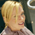 Pixelated close-up of a person with short blonde hair wearing a light-colored collared shirt against a dark background.