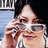 Pixelated close-up of a person with light skin holding a small object near their face, wearing dark sunglasses and a light-colored shirt against a blurred background.