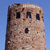 A pixelated, vertically oriented structure resembling a stone or adobe building with multiple rectangular openings against a solid blue background.