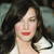 A pixelated close-up of a person with long dark hair, red lips, and light skin, wearing a white top against a blurred background.