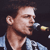 Pixelated close-up of a person singing into a microphone, wearing a light blue shirt against a dark background.