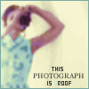 A person in a light blue patterned shirt holds a camera to their face, with text below reading "THIS PHOTOGRAPH IS ROOF" in dark letters.