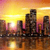 Pixelated cityscape at sunset with buildings reflected in water, warm yellow-orange sky transitioning to pink and purple hues.