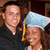 Two people in formal attire, one wearing a graduation cap with a yellow tassel, standing close together against a dark background.