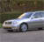 A silver sedan with visible headlights and side windows appears in motion on a road, set against a blurred background of greenery and pavement.