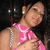 A woman in a pink sleeveless top with white patterns and silver shoulder straps poses with her hand near her chest against a dark background.
