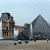 Pixelated static image shows a figure in dark clothing walking left to right past a tall stone tower and a large pyramid-like structure under a light blue sky.