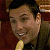 A pixelated close-up of a person with short dark hair, wearing a dark jacket over a light-colored shirt, holding a yellow object near their mouth.