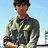 Pixelated image of a person wearing a dark cap, dark jacket with light patches, and light-colored shorts against a light blue background.