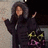 A person in a dark hooded coat with fur trim stands against a textured wall, arms raised in a celebratory pose, wearing a yellow and black patterned bag with pink accents.