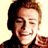 Pixelated static image of a person with light brown hair and closed eyes smiling, wearing dark clothing against a pale yellow background.