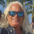 Pixelated portrait of a person with light-colored hair wearing dark sunglasses and a dark collared shirt against a blurred outdoor background.