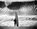 Black-and-white photograph of a surfer standing in shallow water holding a surfboard, facing a large breaking wave under an overcast sky.
