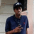 Pixelated image of a person wearing dark cap and blue shirt holding an object, standing against light-colored wall with darker vertical element.