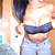 A person in a dark strapless top and light blue shorts poses with hands near shoulders, showing chest area against blurred background.