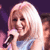 Static pixelated image of a person with long blonde hair holding a microphone against a dark purple background.