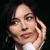 Pixelated close-up of a person with light skin tone, dark hair, and a white garment, hand near face against dark background.