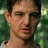 Pixelated close-up of a person's face with short dark hair, light skin, and a neutral expression against a green foliage background.