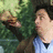 Pixelated image of a person with dark hair and light skin wearing a blue shirt, standing near a tree with green foliage and a light-colored background.