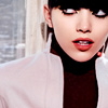 A close-up of a person with dark hair and bangs wearing a white jacket over a dark turtleneck, red lipstick, and red earrings, with a blurred background.