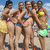 Four women in swimwear pose on a beach with blue sky and clouds, one holding a green frisbee-like object.