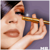 A woman with dark hair applies makeup using a gold applicator near her eye, with 'MB' text visible in the corner.