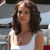 A pixelated image of a person with shoulder-length brown hair wearing a white sleeveless top, set against a blurred background with light and dark tones.