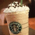 Pixelated image of a light brown beverage in a cup with white foam and dark sprinkles on top, featuring a circular green logo.