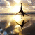 Silhouetted figures of two dancers in mid-pose against a bright, sunlit sky with layered clouds and water reflections.