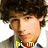 A pixelated image of a person with curly brown hair wearing a dark jacket over a white shirt and red tie, with yellow text at the bottom.