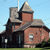 Pixelated image of a multi-story building with brown brick walls, gray stone accents, and a conical roof under a light blue sky.