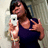 A person in a dark purple tank top holds a white smartphone, making a peace sign with their left hand against a light textured background.
